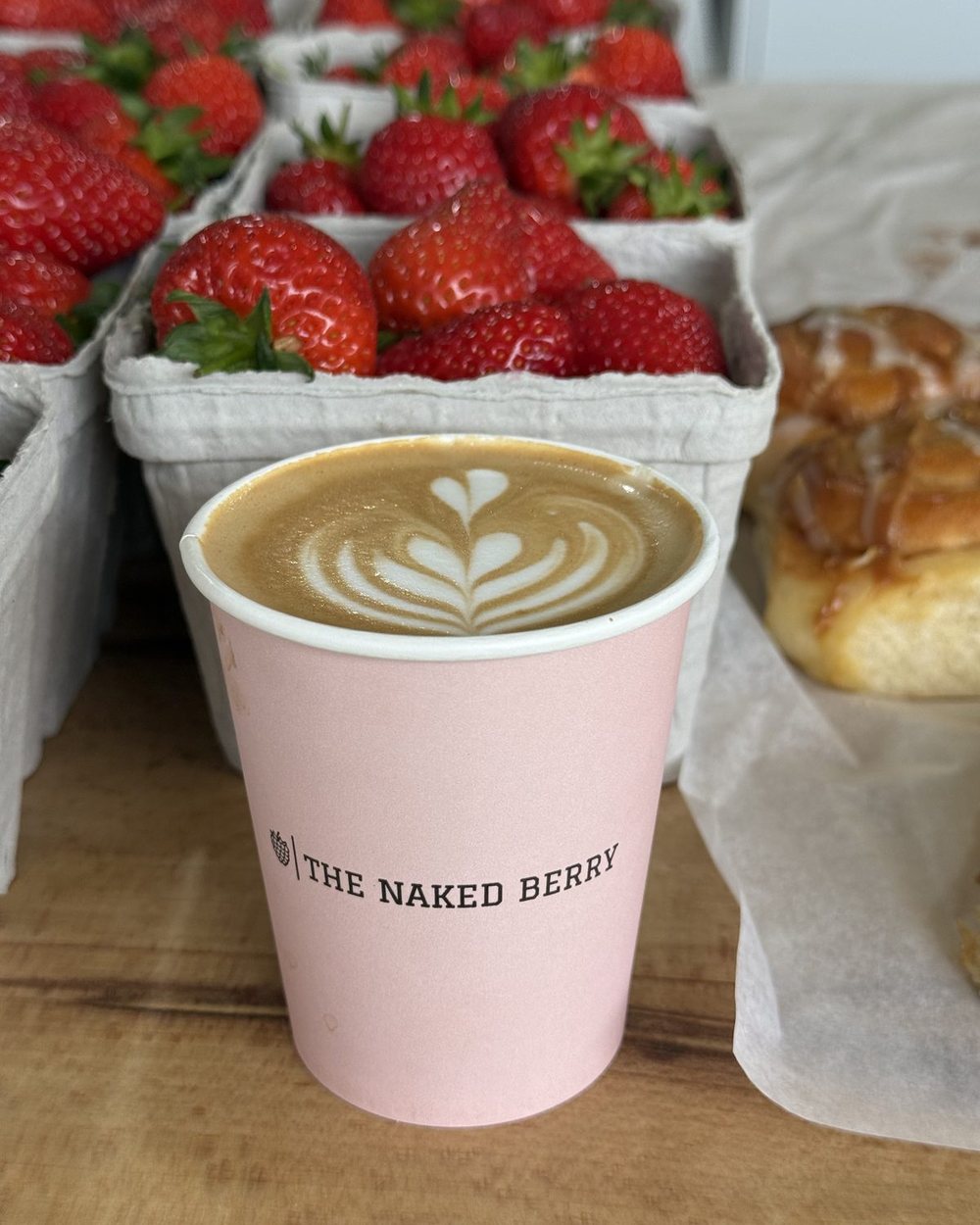A latte with latte art in a pink Naked Berry cup beside fresh strawberries and a pastry